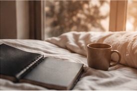 A notebook and warm drink beside a softly lit bed.